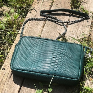 May include: A dark green snakeskin handbag with a silver chain strap. The bag is sitting on a wooden surface.