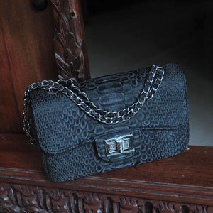 Puede incluir: Un bolso de mano de cuero negro con una correa de cadena plateada y un patrón de cocodrilo texturizado. El bolso tiene un cierre de broche plateado.