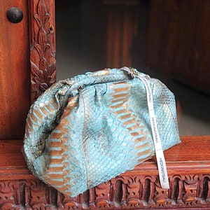 Può includere: Una pochette in pelle di serpente turchese e oro con una chiusura dorata e un'etichetta bianca con la scritta "Hedonistbali".