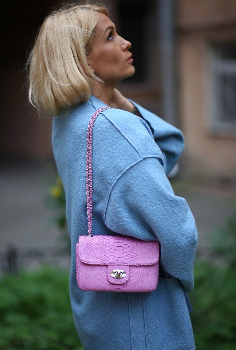 K&ouml;nnte beinhalten: Eine Frau tr&auml;gt einen hellblauen Pullover mit einer rosa Schlangenleder-Schultertasche. Die Tasche hat einen silbernen Verschluss und einen Kettenriemen.
