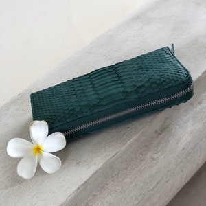 以下が含まれることがあります： 銀色のジッパーが付いた濃い緑色のヘビ革財布。財布は白い表面に白い花と一緒に置かれています。
