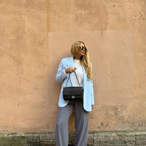 May include: A light blue blazer paired with gray wide-leg pants and white sneakers. A black shoulder bag with a chain strap is worn. The outfit is set against a textured peach-colored wall.