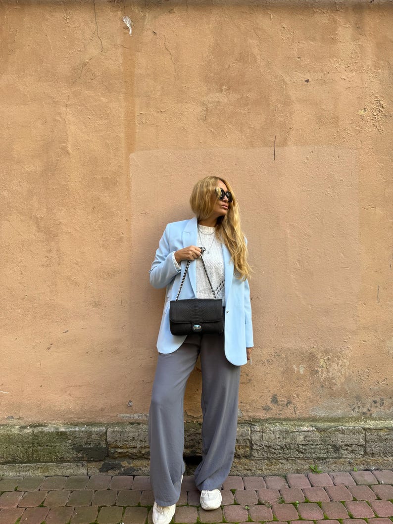 May include: A light blue blazer paired with gray wide-leg pants and white sneakers. A black shoulder bag with a chain strap is worn. The outfit is set against a textured peach-colored wall.