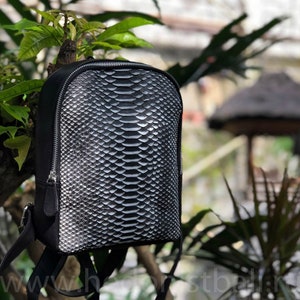 Może przedstawiać: Czarny skórzany plecak ze wzorem skóry węża. Plecak ma zamek błyskawiczny i regulowane paski.