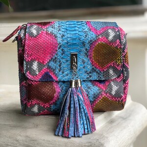 May include: A colorful, patterned handbag with a flap closure. The bag features a vibrant mix of pink, blue, brown, and gray hues, resembling snakeskin. A silver chain and tassel add detail. The bag is sitting on a white surface.