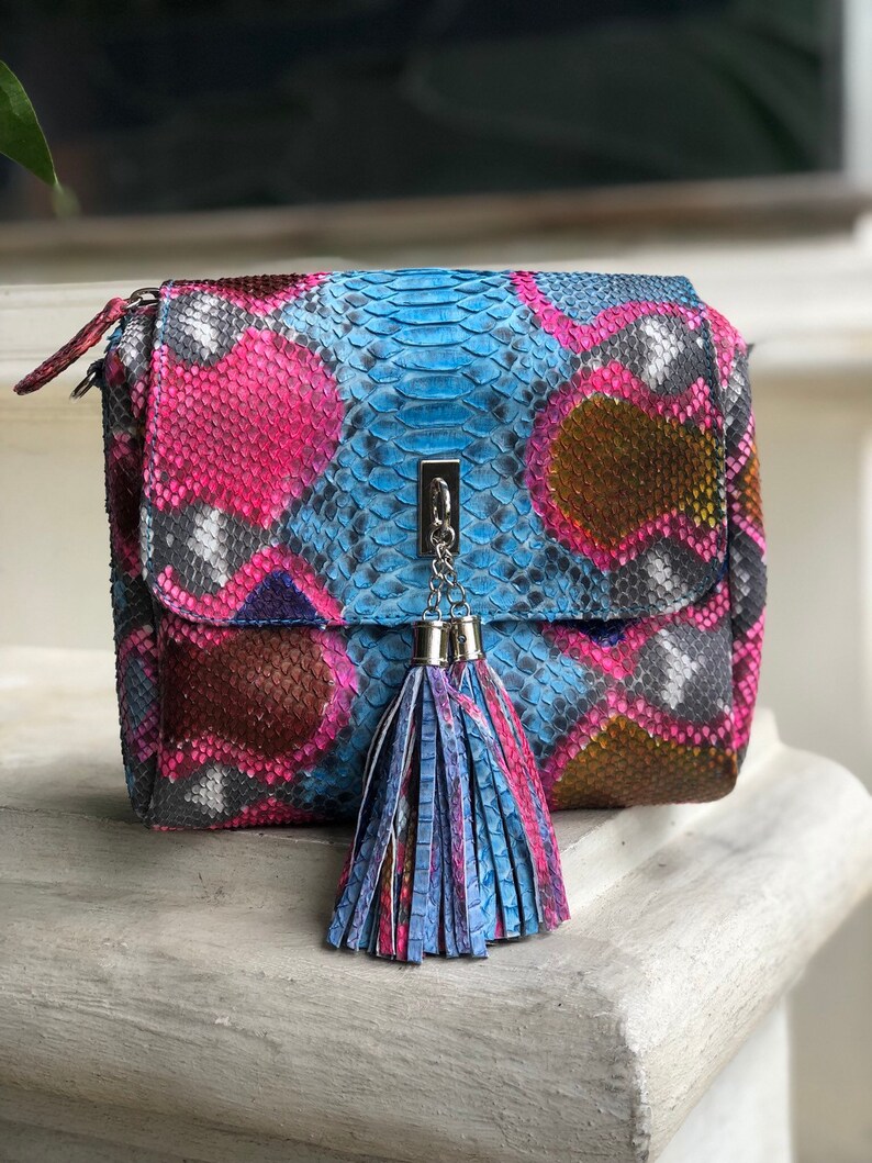 May include: A colorful, patterned handbag with a flap closure. The bag features a vibrant mix of pink, blue, brown, and gray hues, resembling snakeskin. A silver chain and tassel add detail. The bag is sitting on a white surface.