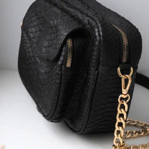 Puede incluir: Bolso bandolera de cuero negro con una correa de cadena dorada y un patrón de piel de serpiente texturizado. El bolso tiene dos compartimentos con cremallera y un cierre de solapa.