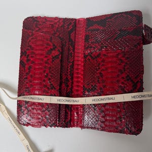 Może przedstawiać: Czerwony i czarny portfel ze skóry węża, otwarty, aby odsłonić wewnętrzne kieszenie. Portfel ma teksturowaną powierzchnię i pasek z napisem "HEDONISTBALI".