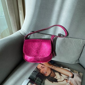 Könnte beinhalten: Eine hellrosa, strukturierte Lederschultertasche mit Klappenverschluss. Die Tasche liegt auf einem grauen Stuhl mit einem Magazin darauf.