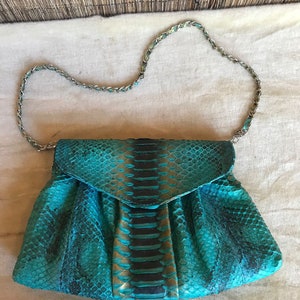Könnte beinhalten: Eine tealblaue Clutch aus Schlangenleder mit einem silbernen Kettenriemen.