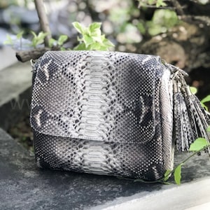 May include: A white and black snakeskin crossbody bag with a tassel. The bag has a flap closure and a shoulder strap.