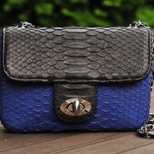 以下が含まれることがあります： 銀色の留め金とチェーンストラップが付いた、黒と青のヘビ革製の小さなハンドバッグです。