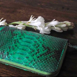 以下が含まれることがあります： ジッパー付きの緑色のヘビ革財布。財布は木製の表面に白い花と一緒に置かれています。
