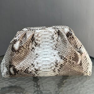 May include: A clutch bag with a snakeskin pattern. The bag is mainly white with brown and black markings. It has a gathered top and a rectangular shape. The snakeskin texture is clearly visible.