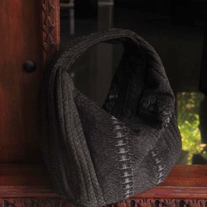 May include: A dark grey handbag with a distinctive textured pattern, possibly resembling scales. The bag has a rounded shape with a single handle. It rests on a wooden surface with carved details, creating a contrast of textures.