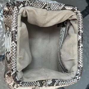 May include: Open, empty handbag with a snakeskin pattern in shades of gray, white, and black. The interior is a light beige fabric. The bag has a silver-colored clasp and zipper.