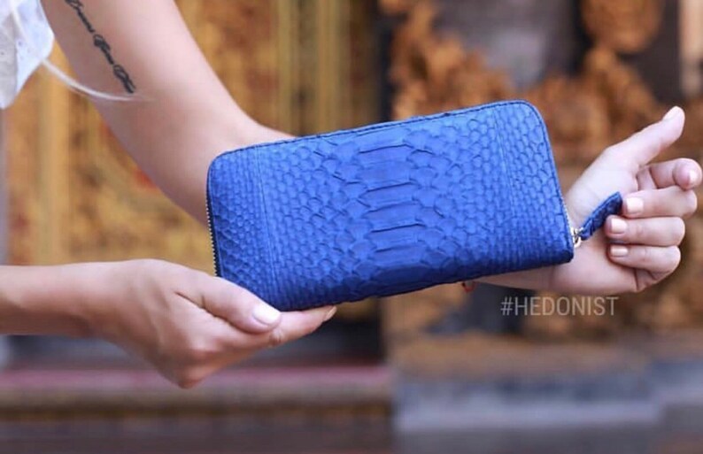 May include: A blue snakeskin wallet with a zipper closure. The wallet is held in a person's hand. #HEDONIST