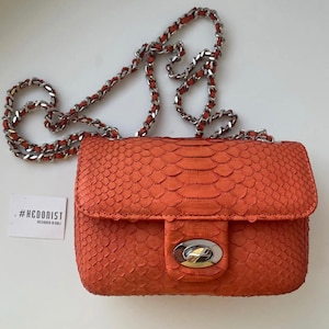 Könnte beinhalten: Eine kleine orangefarbene Schlangenledertasche mit einem silbernen Kettenriemen und einem goldfarbenen Verschluss. Die Tasche trägt den Text "#HEDONIST" und "DESIGNED IN BALI" auf einem weißen Etikett.