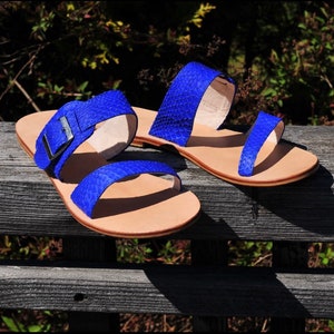 May include: A pair of blue sandals with a textured pattern. The sandals have two straps across the top of the foot and a buckle closure on one. The soles are a light tan color. The sandals are displayed on a weathered wooden bench.