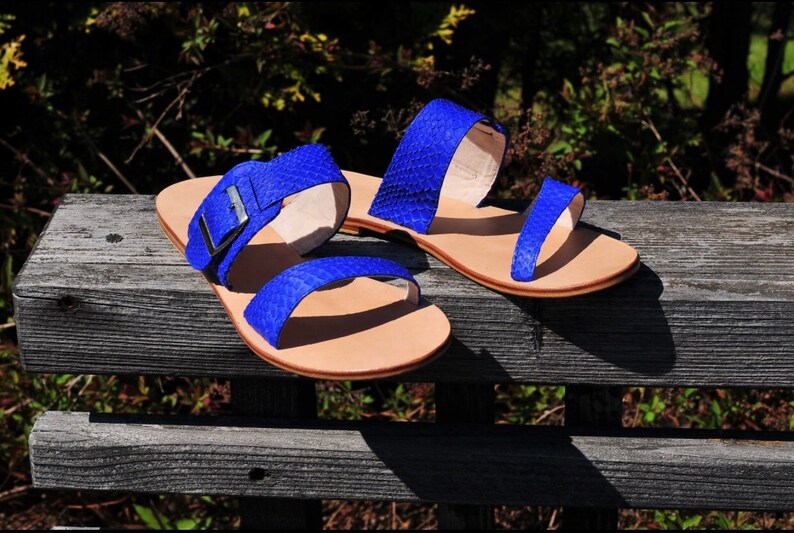 May include: A pair of blue sandals with a textured pattern. The sandals have two straps across the top of the foot and a buckle closure on one. The soles are a light tan color. The sandals are displayed on a weathered wooden bench.