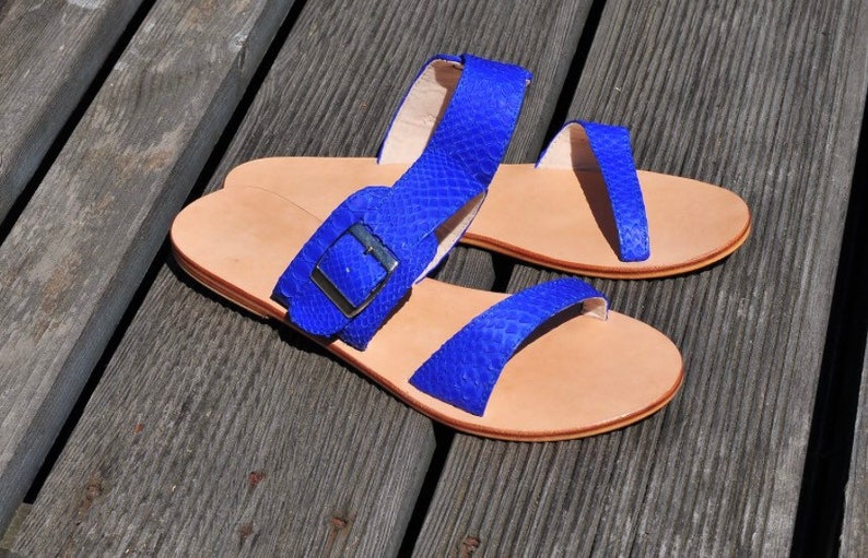 May include: A pair of blue leather sandals with a snakeskin texture. The sandals have a light brown sole and a silver buckle. The sandals are displayed on a wooden surface.