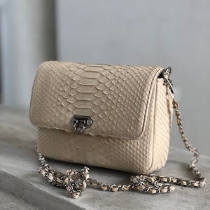 May include: A cream-colored, rectangular handbag with a flap closure and a silver chain strap. The bag is made of a snakeskin-like material, featuring a textured pattern. The clasp is silver, and the bag appears to be sitting on a gray surface.
