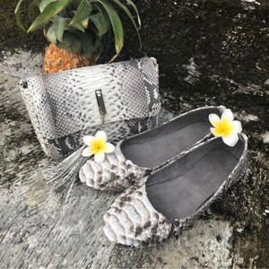 May include: A matching set of accessories featuring a clutch and a pair of flats, both crafted from a snakeskin-like material in shades of gray and white. The clutch has a silver clasp and tassel. The flats are adorned with a yellow and white flower.