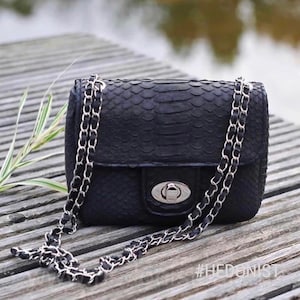 Könnte beinhalten: Eine schwarze Ledertasche mit einer silbernen Kettenkette und einem silbernen Verschluss. Die Tasche hat ein strukturiertes, schlangenartiges Muster. #HEDONIST