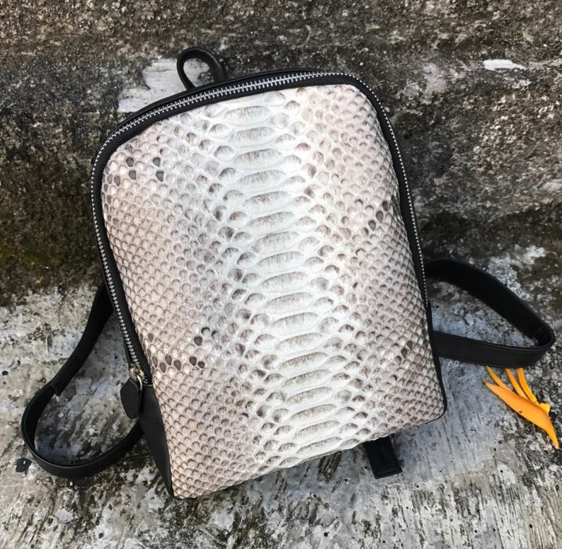 以下が含まれることがあります： 白とグレーのヘビ革のバックパックで、黒いストラップとジッパーが付いています。