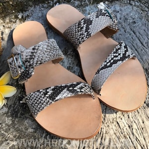 May include: A pair of grey and white snakeskin sandals with a buckle fastening. The sandals have a flat sole and are made of leather.