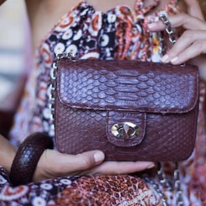 May include: A brown snakeskin purse with a silver clasp and a chain strap. The purse is held in the hand of a person wearing a patterned dress.