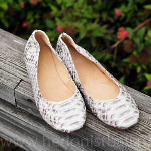 May include: A pair of white and gray snakeskin-patterned ballet flats. The shoes are made of a textured material and have a rounded toe. The interior of the shoe is a light tan color. The flats are displayed on a wooden surface.