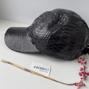 以下が含まれることがあります： ヘビ柄の黒革製のベースボールキャップ。キャップはカーブしたつばと、"designed in bali"というテキストが書かれた#HEDONISTのラベルが付いています。