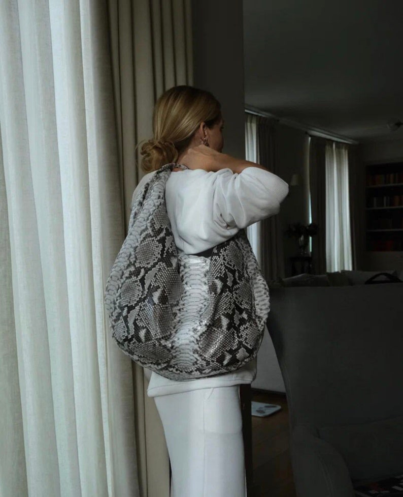 May include: A woman wearing a white sweater and white skirt is holding a large, gray and black snakeskin handbag.