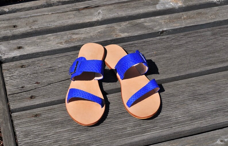 May include: A pair of open-toe sandals with a light brown sole and bright blue straps. The straps are textured and feature a buckle closure. The sandals are displayed on a weathered wooden surface.