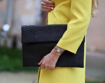 May include: A woman is holding a black snakeskin clutch purse with a fold-over flap. The purse is closed and has a silver watch on the wrist of the hand holding the purse.