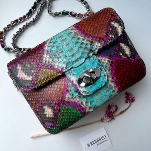 Op de afbeelding: Een klein, veelkleurig schlangenleren handtasje met een zilveren kettingband. De tas heeft een klep sluiting met een zilveren sluiting. De tas ligt op een wit oppervlak met een klein label waarop staat "#HEDONIST DESIGNED IN BALI".