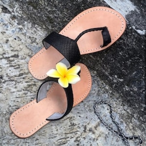 Op de afbeelding: Zwarte leren sandalen met een teenring en een gele bloemversiering. De sandalen hebben een bruine leren zool.