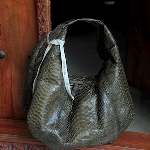 Puede incluir: Un bolso de mano de piel de serpiente verde con una etiqueta blanca que dice "HEDONISTIC TRAIL".