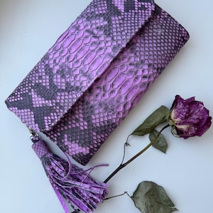 May include: A purple snakeskin wallet with a tassel. The wallet is folded closed and has a textured surface.