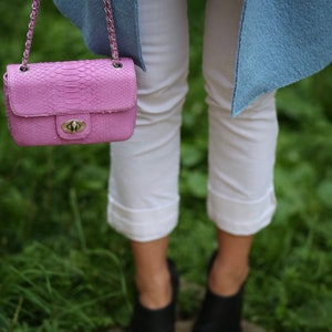 K&ouml;nnte beinhalten: Eine rosa Schlangenleder-Handtasche mit einer silbernen Kettenkette und einem silbernen Schloss. Die Tasche wird von einer Person getragen, die eine wei&szlig;e Hose und eine hellblaue Jacke tr&auml;gt.