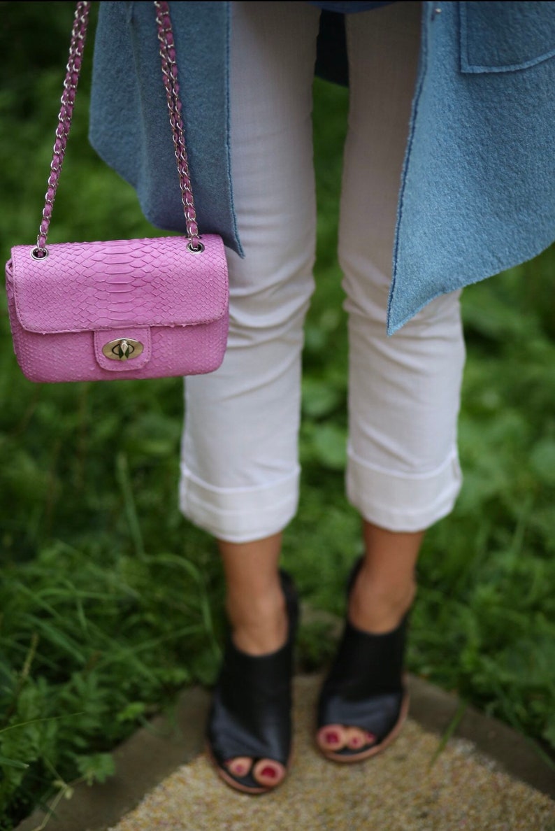 K&ouml;nnte beinhalten: Eine rosa Schlangenleder-Handtasche mit einer silbernen Kettenkette und einem silbernen Schloss. Die Tasche wird von einer Person getragen, die eine wei&szlig;e Hose und eine hellblaue Jacke tr&auml;gt.