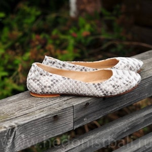 May include: A pair of white and gray snakeskin-patterned ballet flats. The shoes are made of a textured material and have a rounded toe. The soles are a light brown color. The shoes are displayed on a weathered wooden surface.