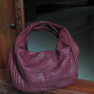 Peut inclure: Un sac à main en cuir bordeaux avec un motif en peau de serpent. Le sac a une seule bandoulière et une large ouverture.