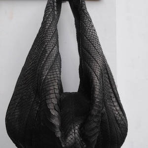 May include: A black leather handbag with a snakeskin pattern. The bag has a single strap and is hanging from a wooden hook.