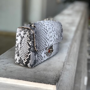 Può includere: Borsa rettangolare con motivo a pelle di serpente grigio e bianco, con tracolla a catena argentata. La borsa ha una chiusura a patta con una piccola chiusura decorativa. Poggia su una superficie grigia.