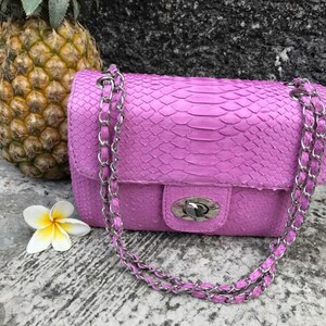 K&ouml;nnte beinhalten: Eine kleine, helllila Handtasche mit einem silbernen Kettenriemen. Die Tasche ist aus Schlangenleder und hat einen Klappverschluss mit einem silbernen Verschluss.