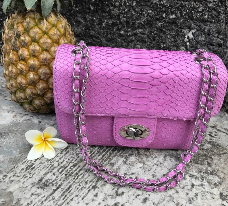 K&ouml;nnte beinhalten: Eine kleine, helllila Handtasche mit einem silbernen Kettenriemen. Die Tasche ist aus Schlangenleder und hat einen Klappverschluss mit einem silbernen Verschluss.