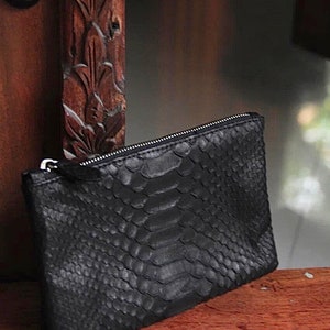 Może przedstawiać: Czarna kopert&oacute;wka ze sk&oacute;ry węża z srebrnym zamkiem błyskawicznym.