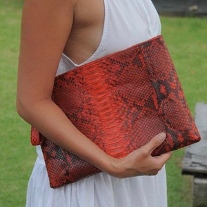 Może przedstawiać: Czerwono-czarna kopertówka ze skóry węża. Kopertówka ma fakturowaną powierzchnię i jest trzymana przez rękę.